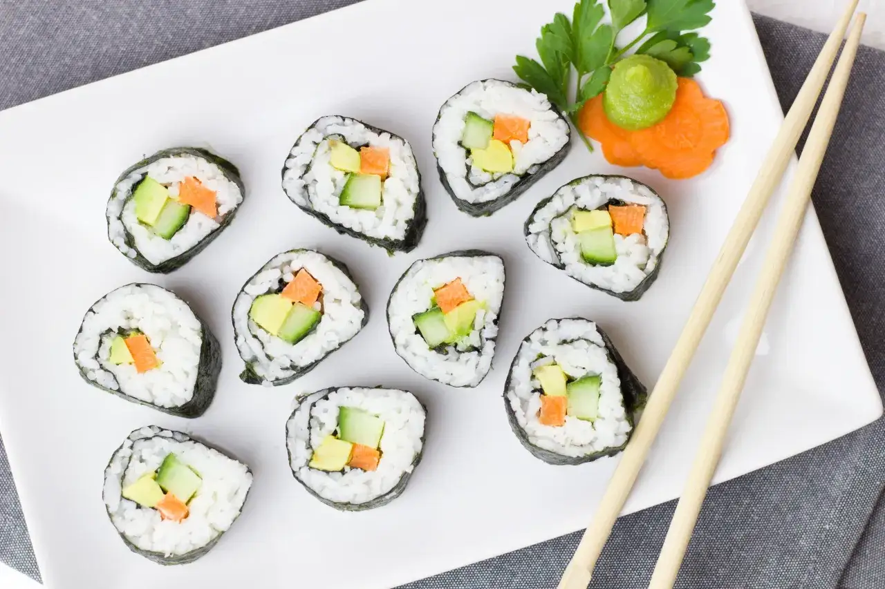 Roladki sushi z awokado, ogórkiem i marchewką. Obok wasabi i ozdobna marchewka. Pałeczki czekają na rybę do sushi.