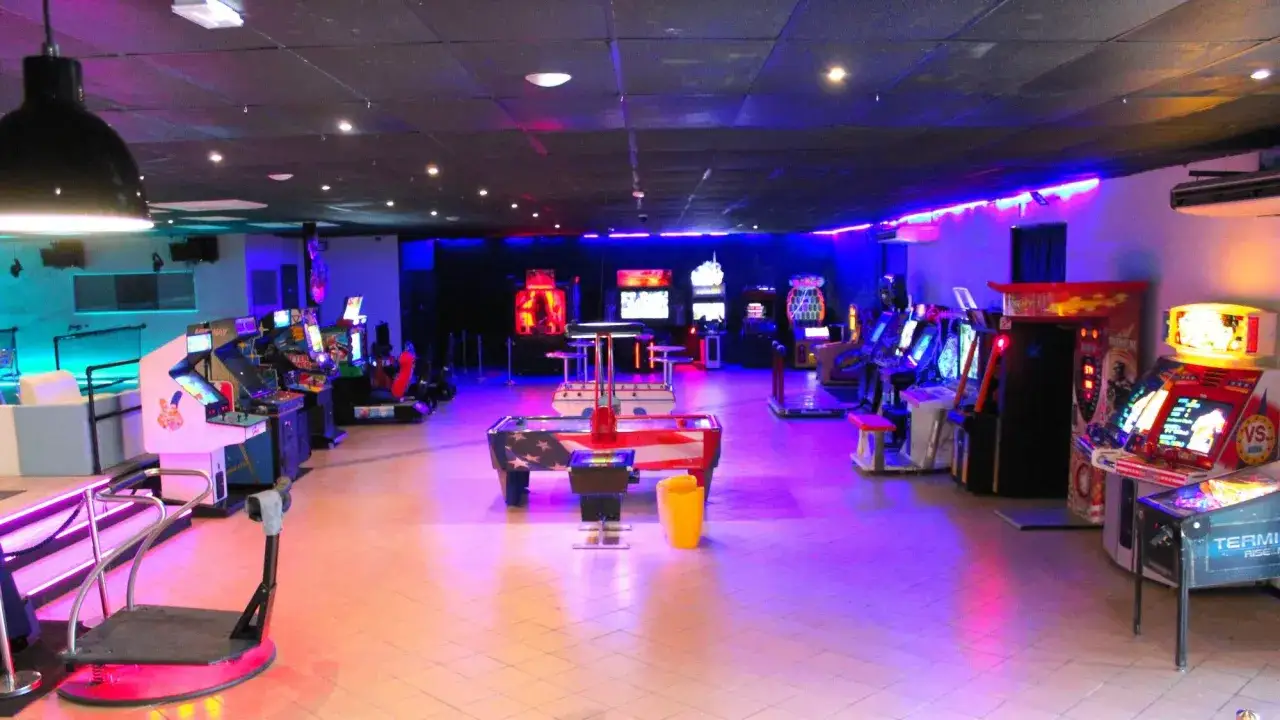 Salle de jeux vidéo illuminée de néons bleus et roses, avec des bornes d'arcade et un baby-foot. L'ambiance rappelle l'**elyzion orange** des soirées d'été.