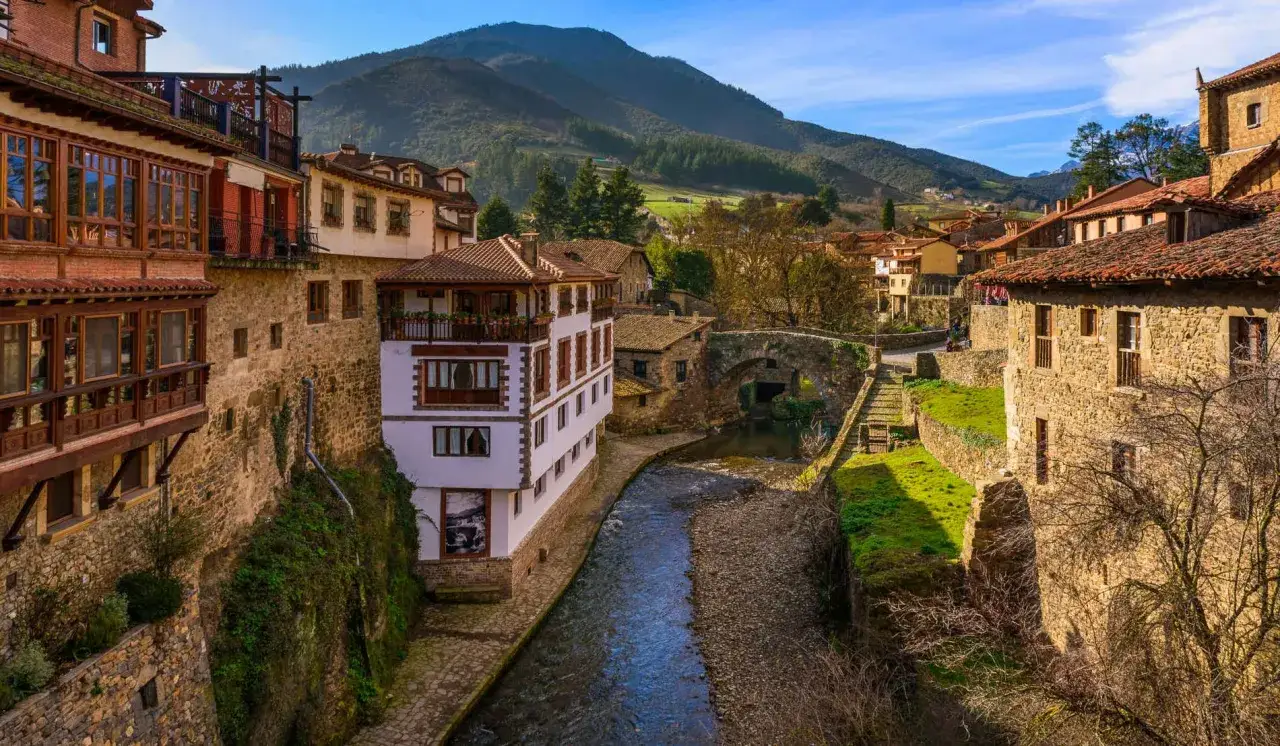 Un r&iacute;o atraviesa un pintoresco pueblo con casas de piedra y balcones de madera, un puente antiguo y monta&ntilde;as al fondo. &iexcl;Pueblos bonitos cerca de Puente Viesgo!