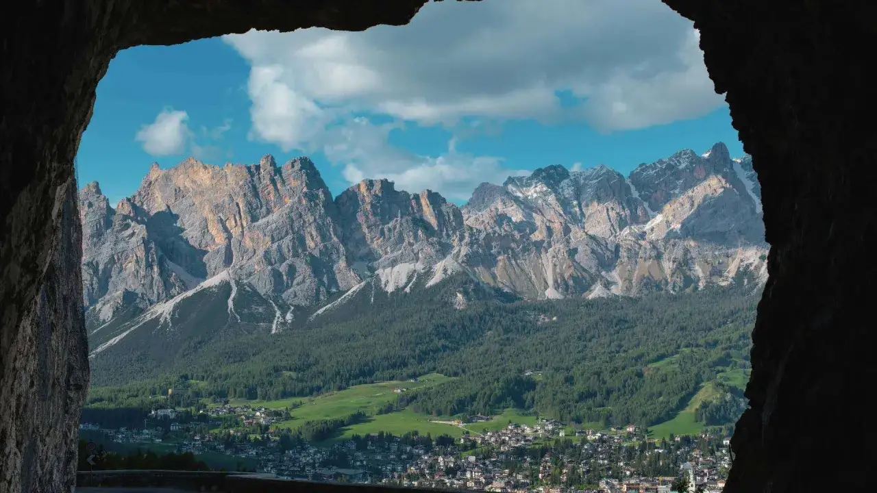 Vista de **Milano Cortina** desde una cueva, con imponentes montañas rocosas, bosques verdes y un pueblo pintoresco.
