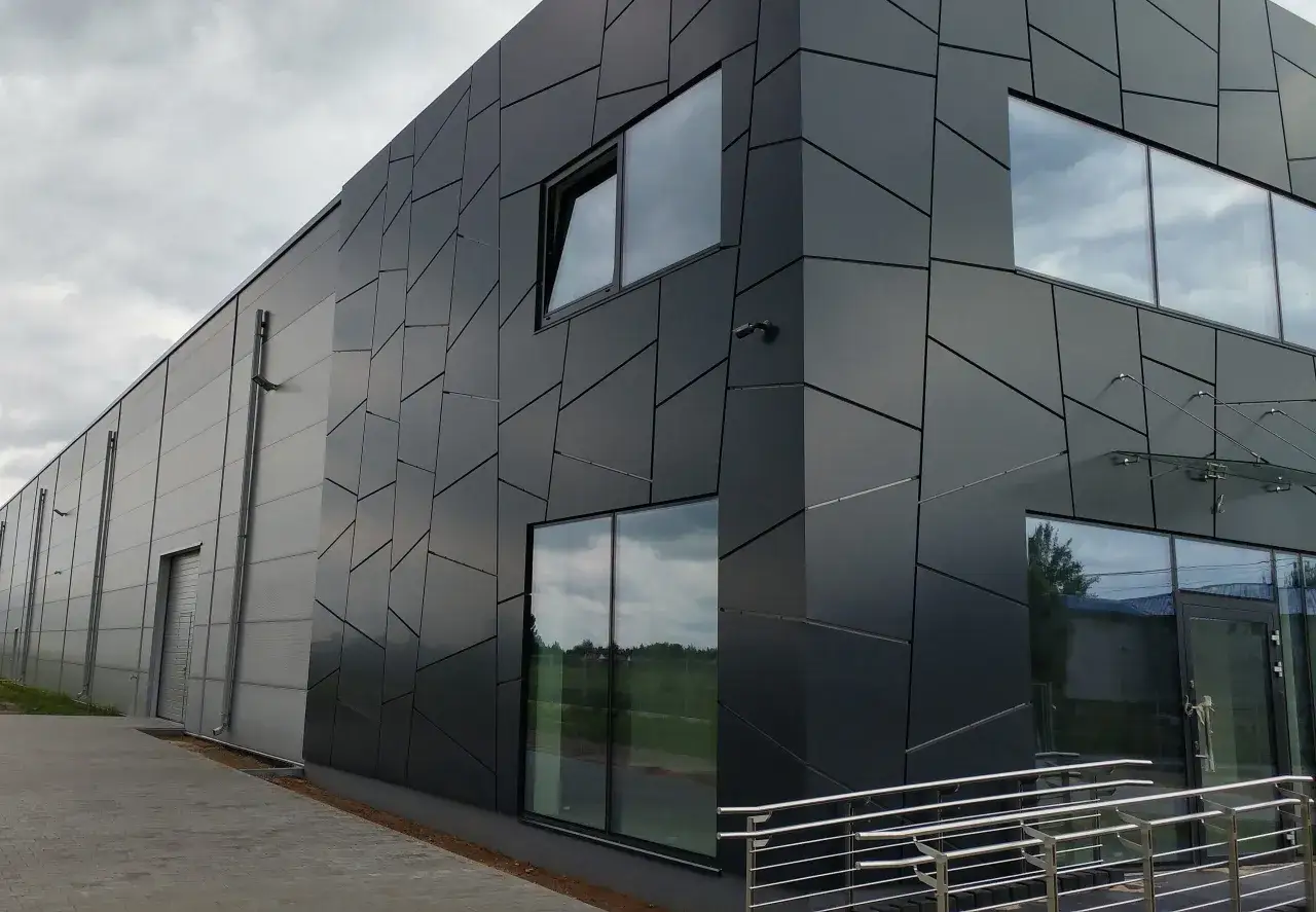 Nowoczesny budynek z czarną, geometryczną elewacją, oknami i drzwiami. Widać fragment chodnika i zieleni. To część większego aglomeratu.