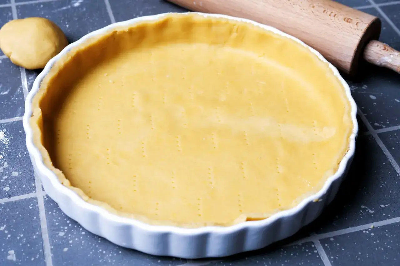 Ein vorbereiteter quiche teig liegt in einer weißen Tarteform. Daneben ein Nudelholz und ein Teigball.