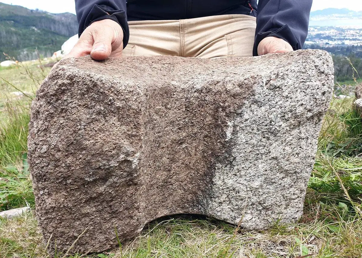 Un hombre sostiene una piedra tallada, vestigio del castro de Chandebrito, con un paisaje monta&ntilde;oso al fondo.