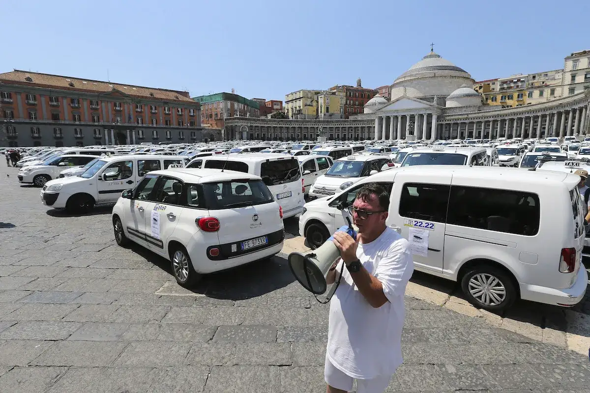 Un uomo con un megafono parla a una folla di taxi bianchi parcheggiati, forse per discutere quanto guadagnano i tassisti.