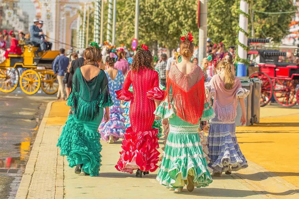 Kobiety w barwnych sukniach flamenco idą na Feria de Abril. W tle wóz konny i tłum.