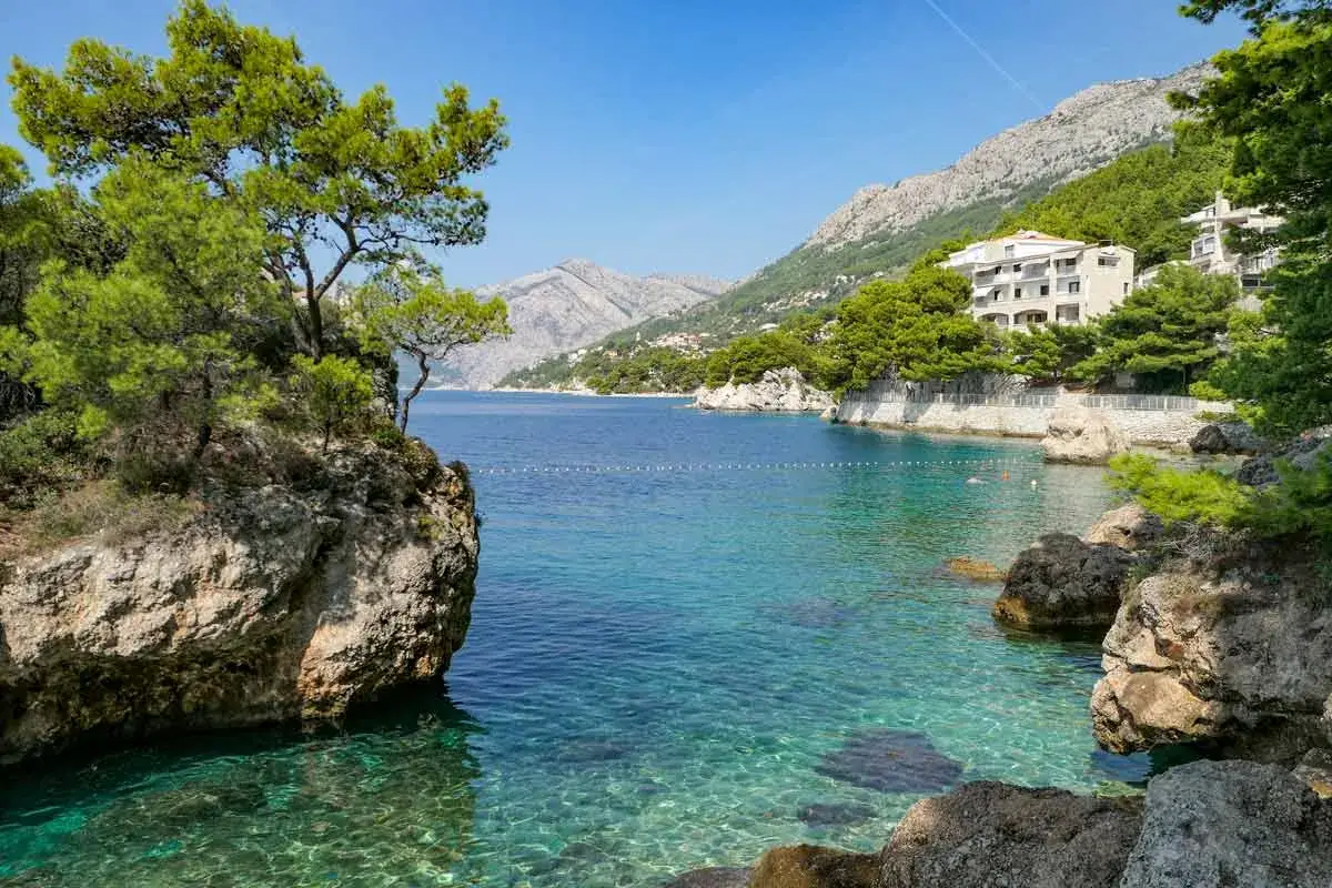 Krystalicznie czysta woda i skaliste wybrzeże na plaży Makarska. W oddali widać góry i biały hotel.