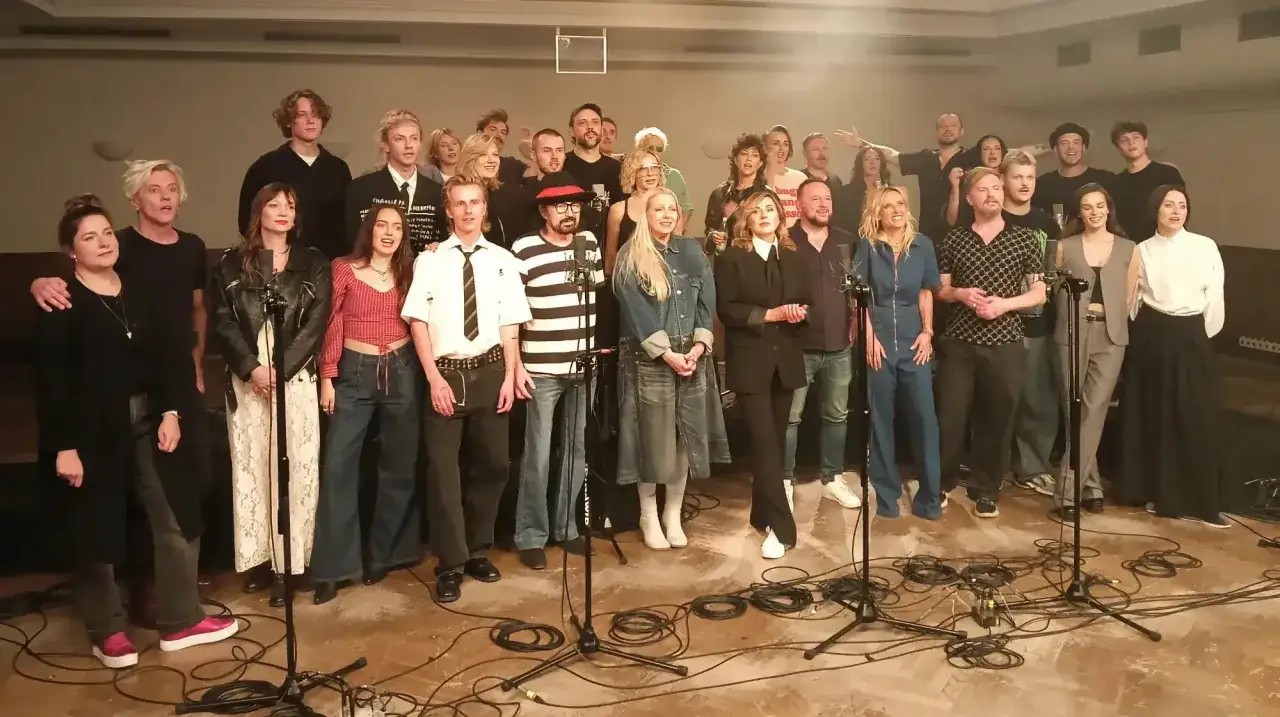 Grupa ludzi w różnym wieku, ubrana w stylowe stroje, stoi przed mikrofonami. Wszyscy wyglądają na gotowych do występu, ale w ich oczach widać pewną rezygnację. Nic naprawdę nic nie pomoże.