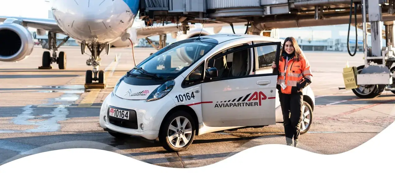 Donna con giacca arancione sorride accanto a un'auto bianca con logo Aviapartner, pronta a lavorare in aeroporto.