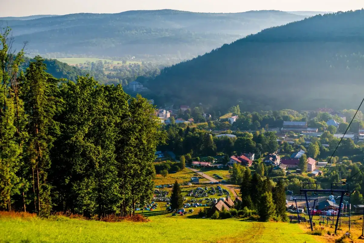 Widok na dolinę z miasteczkiem i polem namiotowym, otoczoną zalesionymi wzgórzami. To urokliwy krajobraz **Małej Pętli Bieszczadzkiej**.