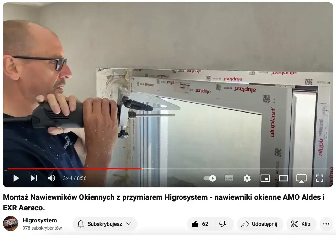 Mężczyzna montuje nawiewniki w oknach, używając specjalnego przyrządu. Widoczne są białe profile okienne z logo