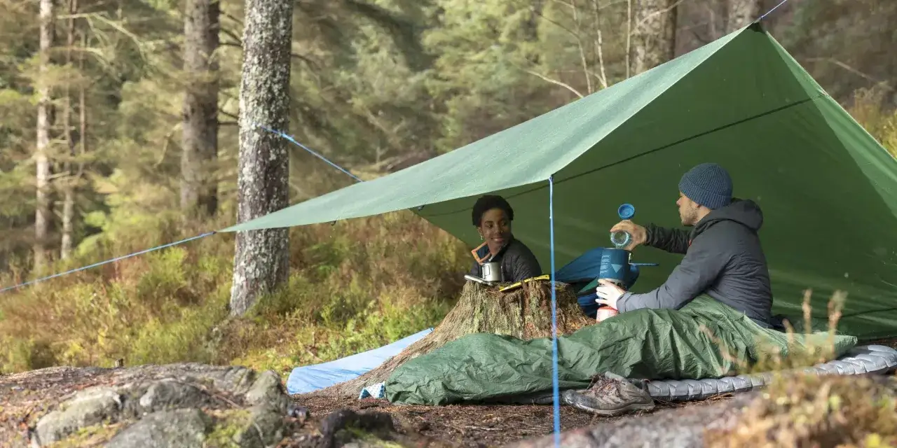 Zwei Camper bereiten unter einem gr&uuml;nen Tarp eine Mahlzeit zu. Das Tarp sch&uuml;tzt sie vor Regen und bietet einen gem&uuml;tlichen Unterschlupf.