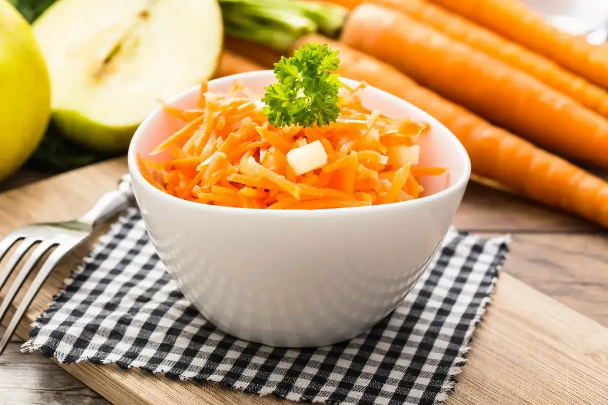 Frischer Karotten-Apfel-Salat, garniert mit Petersilie, neben ganzen Karotten und Apfelhälften.