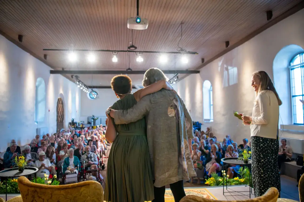 Två kvinnor omfamnar varandra på scenen under bokdagar i Dalsland. Publiken applåderar.