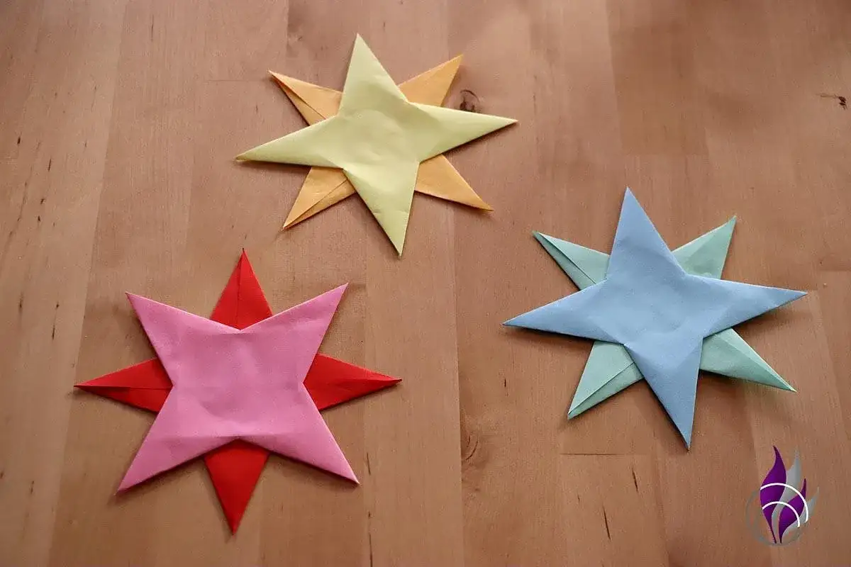 Drei bunte Papiersterne liegen auf einem Holztisch. Sie sind das Ergebnis von **Wurfstern basteln**.