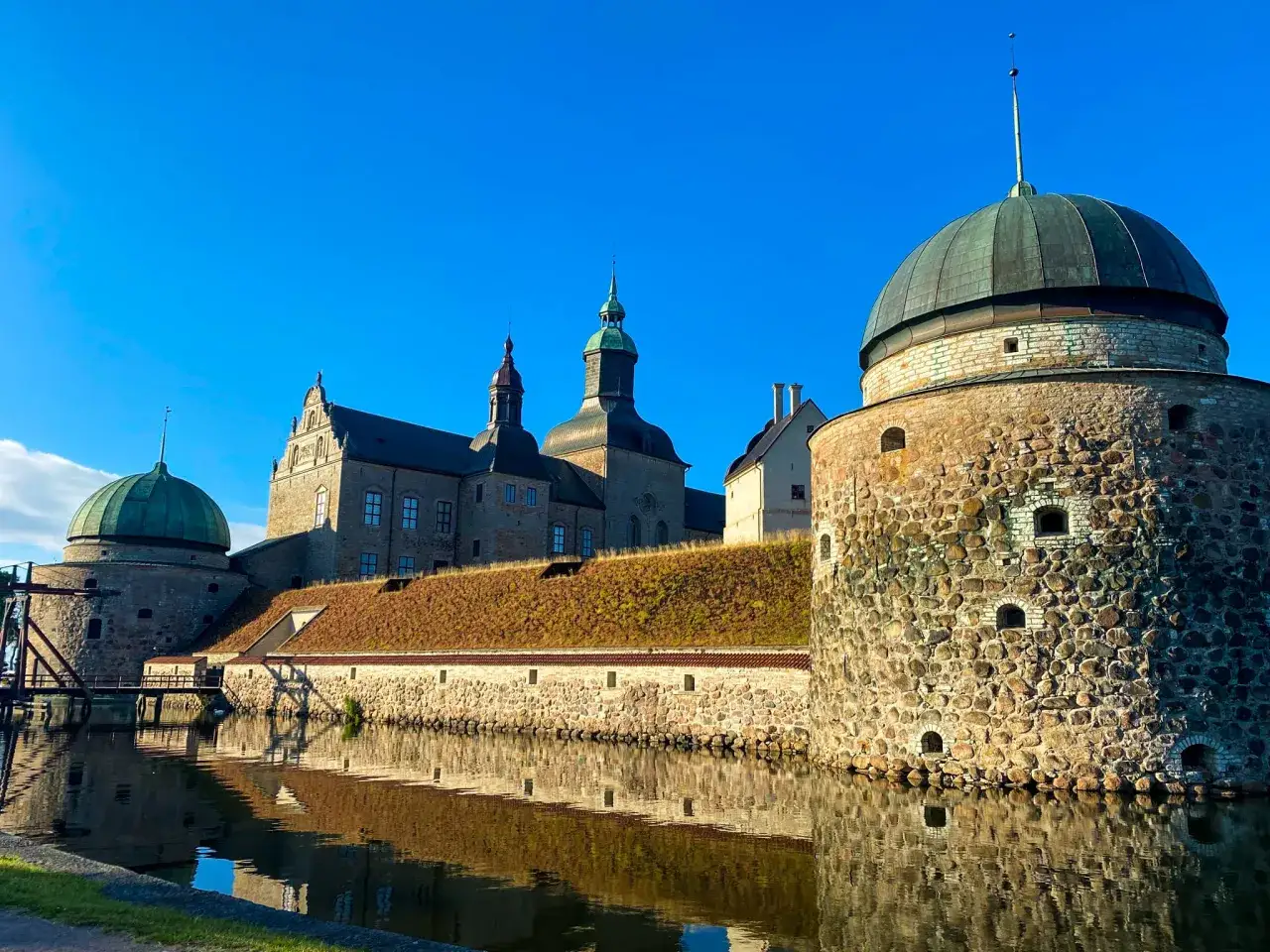 V&auml;lkommen till Vadstena! Imponerande slott med kupoler och vallgrav speglar sig i vattnet under en klarbl&aring; himmel.