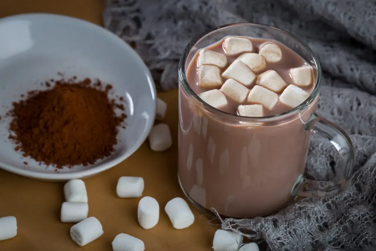 Jak zrobić kakao z wodą? Na talerzyku kakao, obok kubek gorącego napoju z piankami i marshmallow.