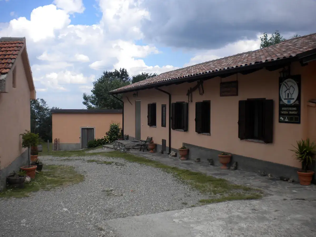Agriturismo Acqua Bianca, un luogo sereno con edifici in pietra e terra, circondato da vegetazione e un cielo nuvoloso.