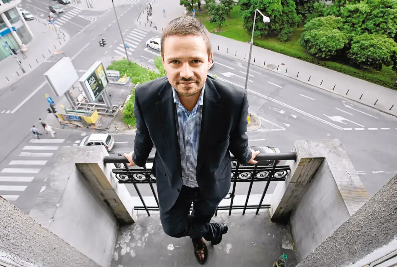 Rafał Trzaskowski, w wieku około 40 lat, stoi na balkonie, patrząc w obiektyw. W tle ruchliwa ulica.