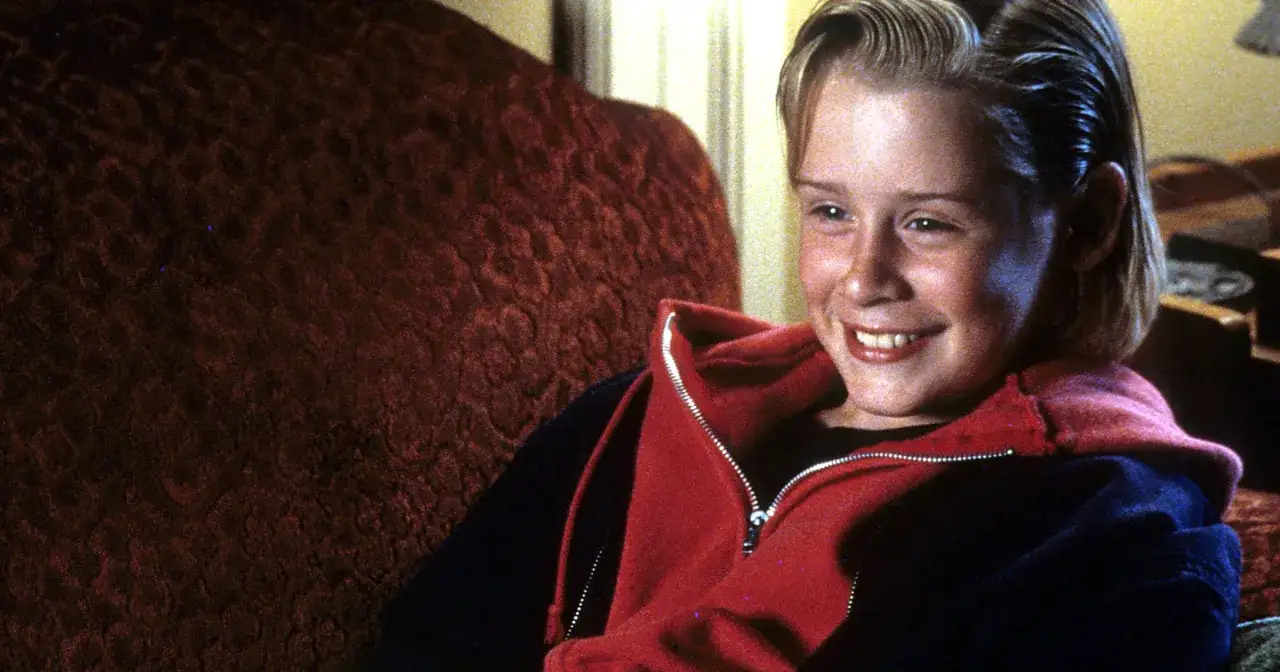 Uśmiechnięty Macaulay Culkin, znany z tego, w jakich filmach grał, siedzi na kanapie w czerwonej bluzie.