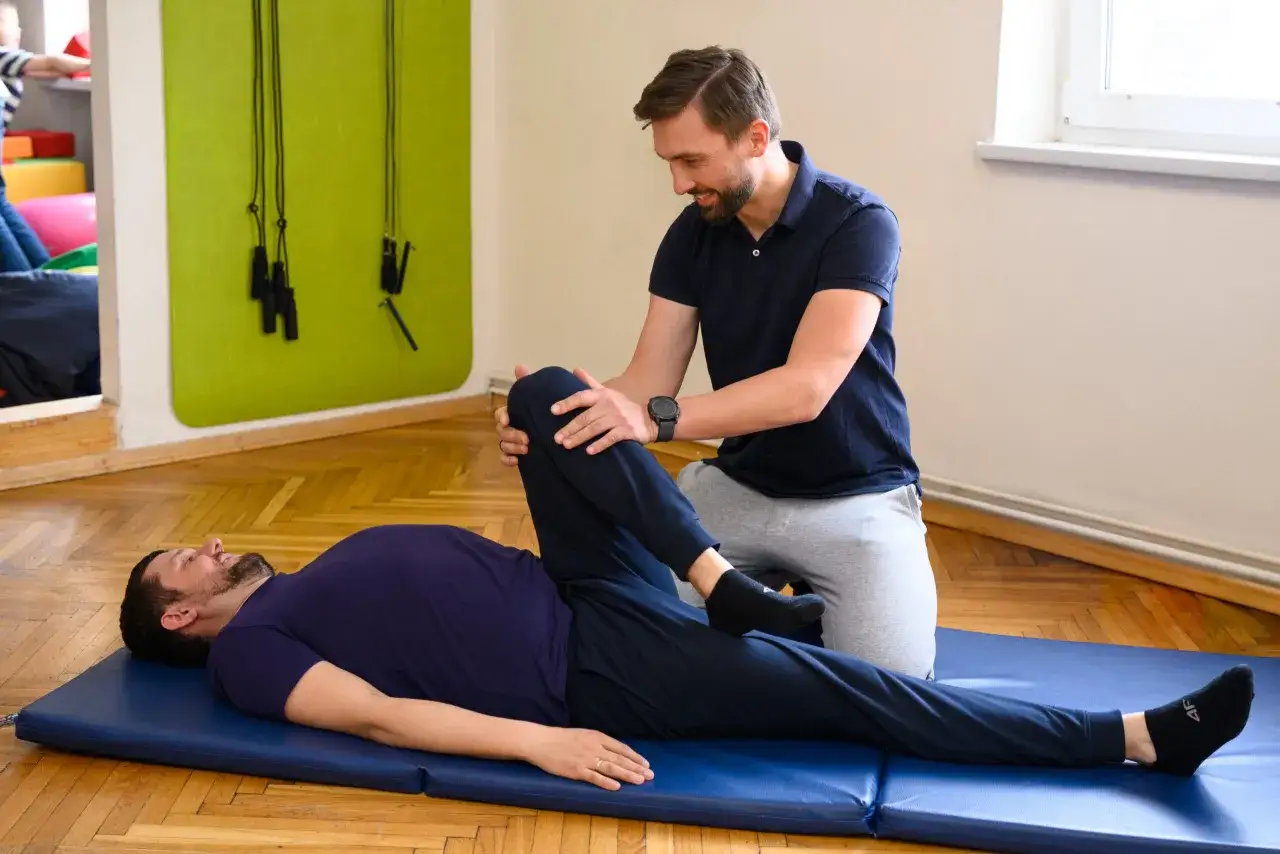 Fizjoterapeuta wykonuje zabiegi rehabilitacyjne, pomagając pacjentowi w ćwiczeniach rozciągających nogę na macie.