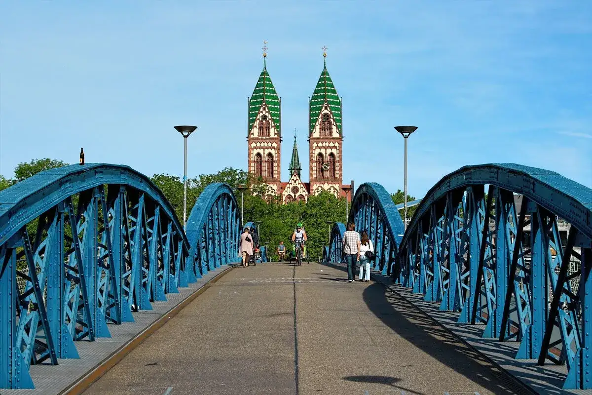 Menschen spazieren und radeln über eine blaue Brücke auf die imposante Kirche zu. So kann man in Freiburg die Stadt erkunden.