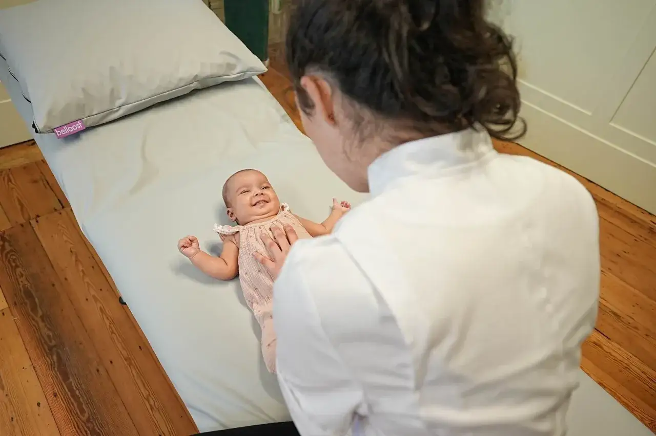 Ein Baby l&auml;chelt auf einer Liege, w&auml;hrend eine Osteopathin sanft seine Brust ber&uuml;hrt. Ein sch&ouml;ner Moment, wann mit Baby zum Osteopathen.