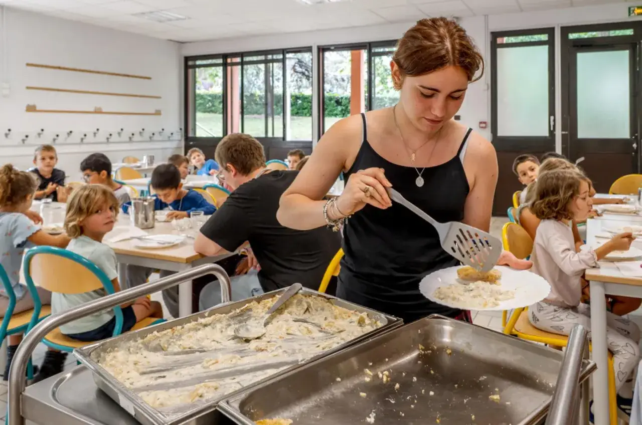 Une jeune femme sert un plat crémeux à des enfants dans une cantine. Fiche technique préparation des repas.