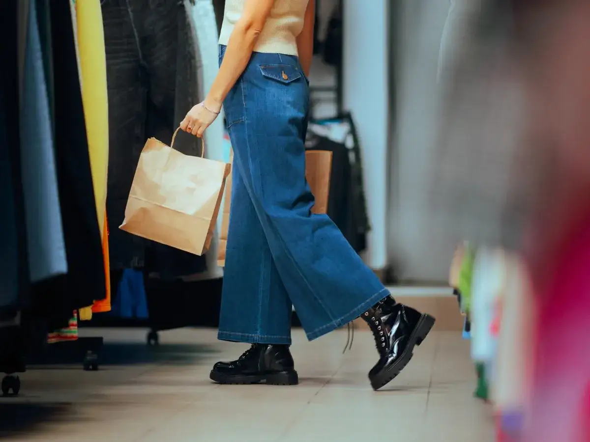Kobieta w jeansach bootcut i czarnych botkach idzie na zakupy, trzymając papierową torbę.