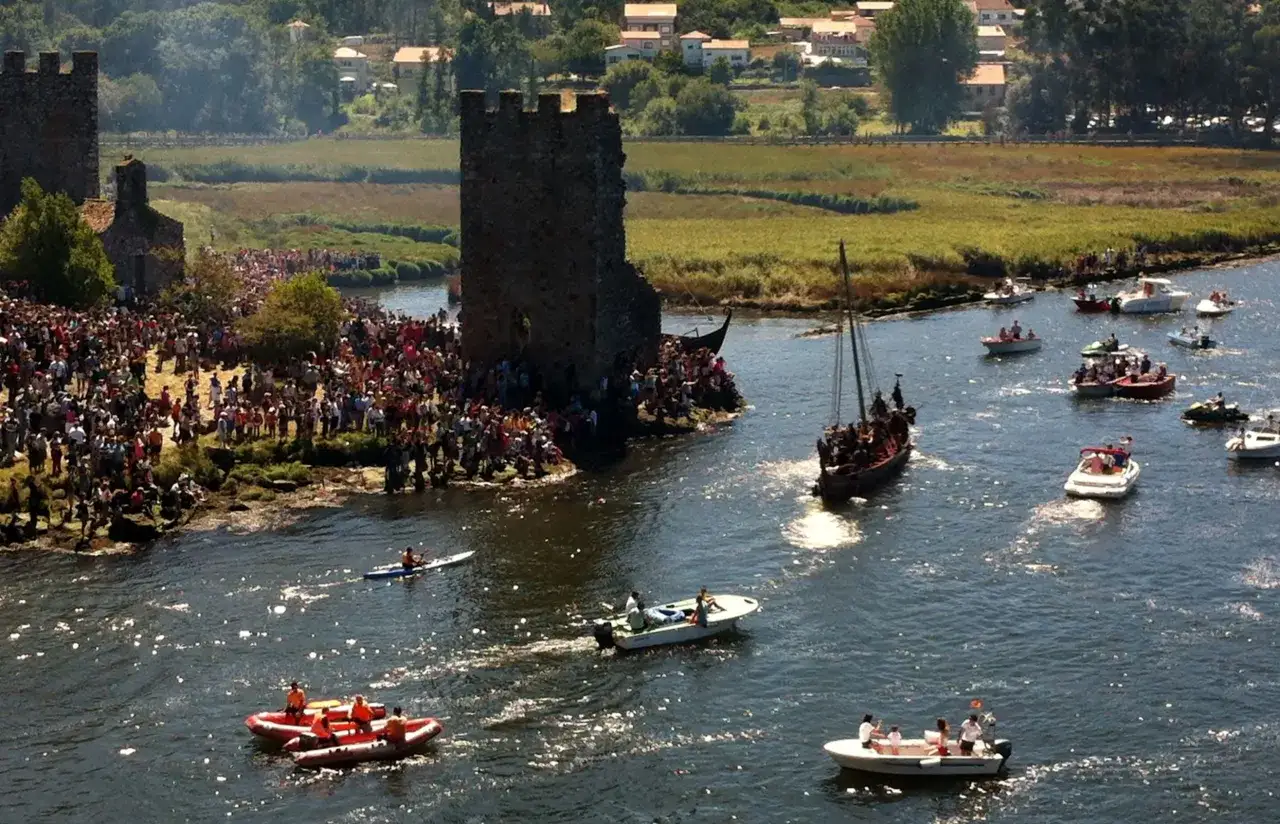 Multitud celebra desembarco vikingo en Catoira, Galicia. Barcos navegan por el r&iacute;o Ulla, con torres medievales de fondo.