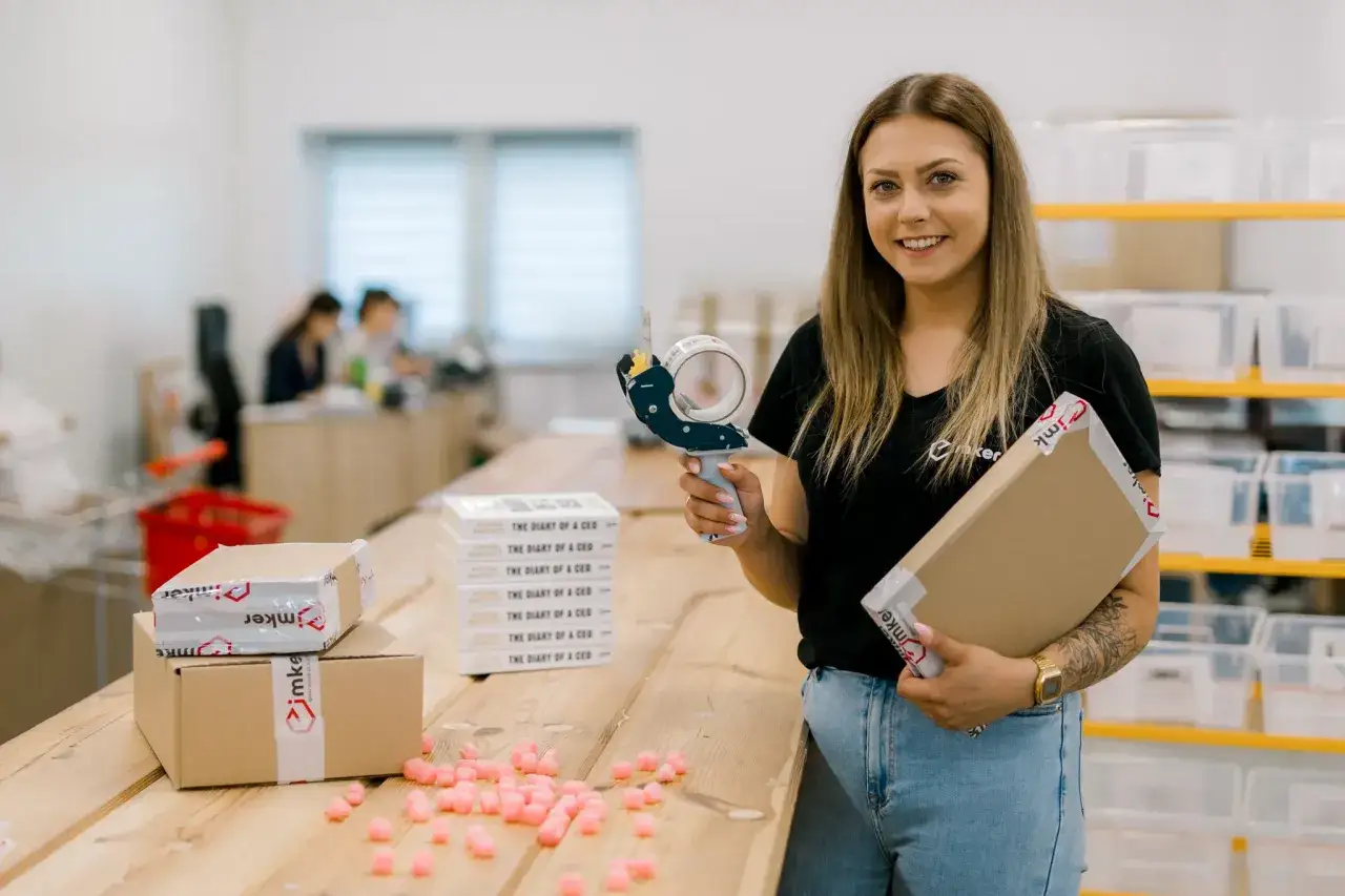 Uśmiechnięta pracownica pakuje paczki do wysyłki, przygotowując zamówienia dla e-commerce.