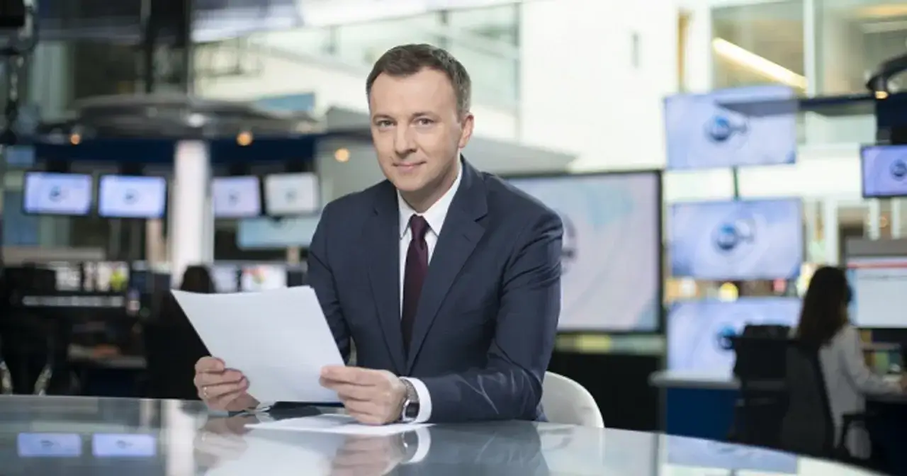 Krzysztof Górlicki w garniturze siedzi przy biurku z dokumentami w studiu telewizyjnym.