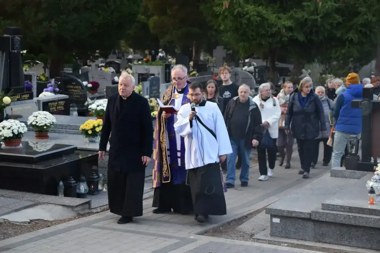 Ksiądz z mikrofonem niesie urnę na pogrzebie, a za nim idzie grupa żałobników przez cmentarz.