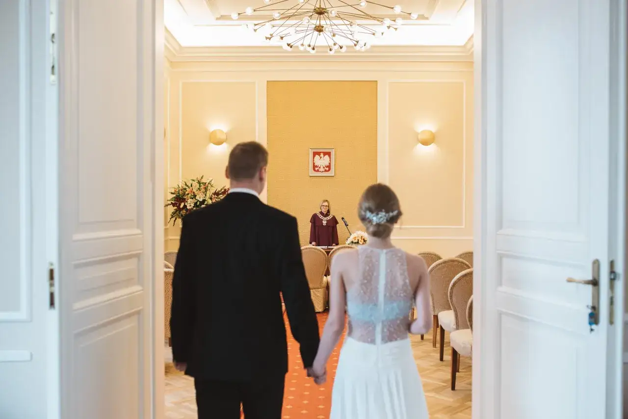 Para młoda idzie na przysięgę małżeńską ślub cywilny. Urzędniczka czeka przy mównicy.