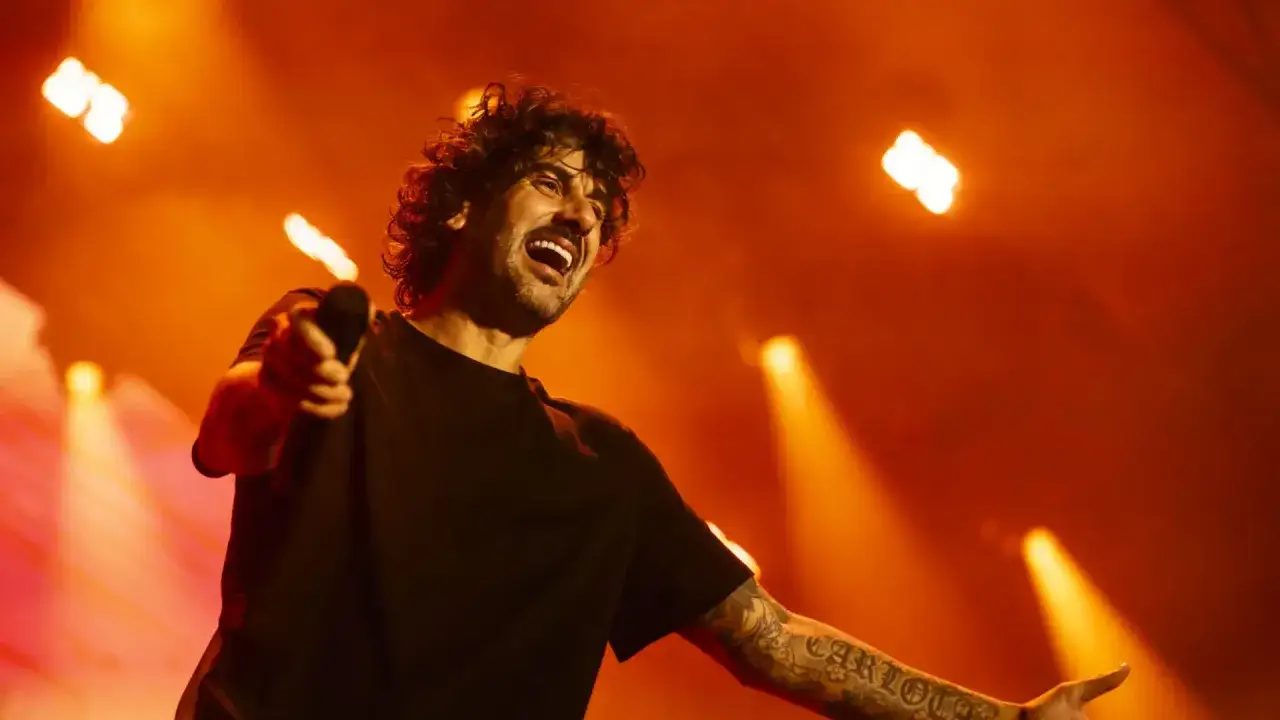 Melendi canta con pasión en su concierto en Zaragoza, iluminado por luces naranjas.