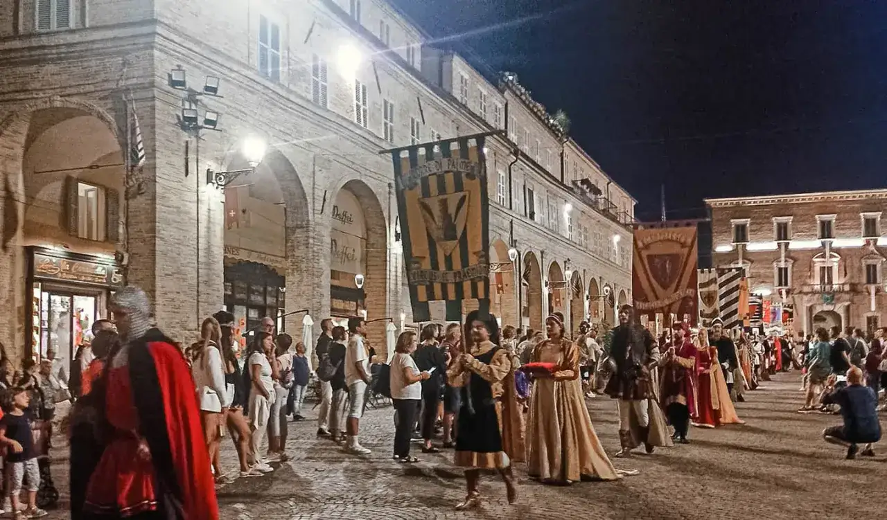 La **cavalcata dell'Assunta a Fermo** sfila di notte tra le arcate. Persone in costume storico portano stendardi colorati, mentre la folla osserva.