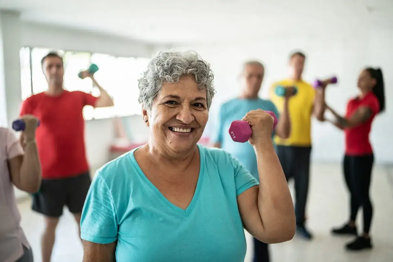 Donna sorridente con manubrio rosa, 50 anni e non sentirli, in palestra con altri che si allenano.