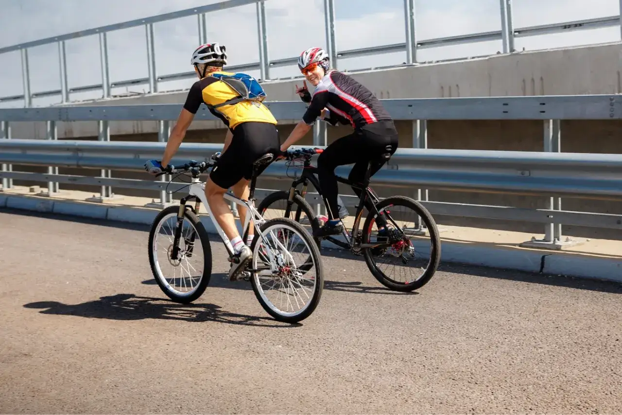 Dw&oacute;ch rowerzyst&oacute;w jedzie obok siebie po moście. Jeden z nich macha do widza.