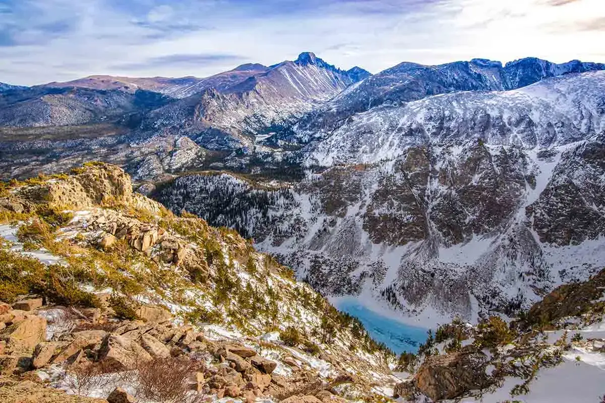 Schneebedeckte Berge und ein gefrorener See entlang des Continental Divide Trail. Felsige H&auml;nge mit Kiefern und blauem Himmel.