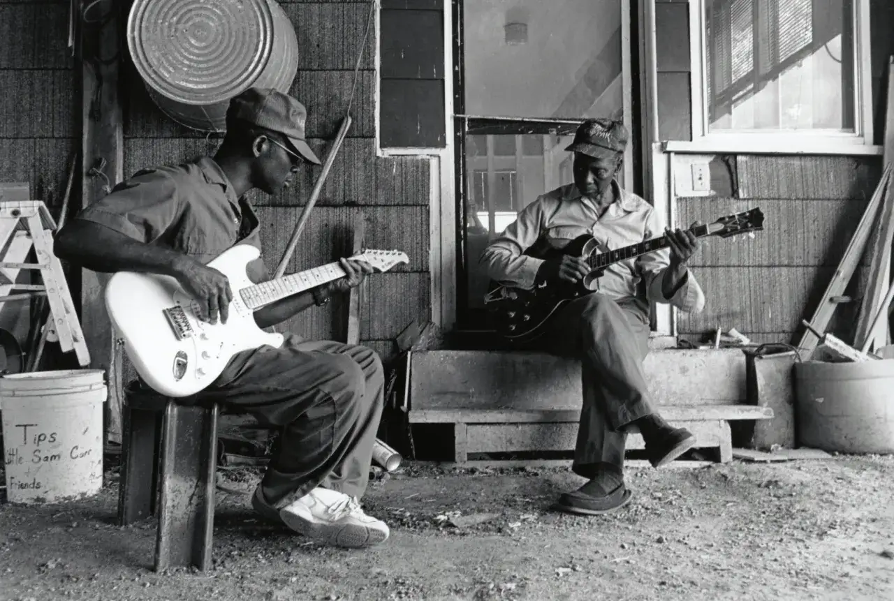 Deux musiciens jouent de la guitare, l'un sur une électrique blanche, l'autre sur une acoustique sombre. Ils incarnent l'ancêtre du blues, une musique née de la terre.