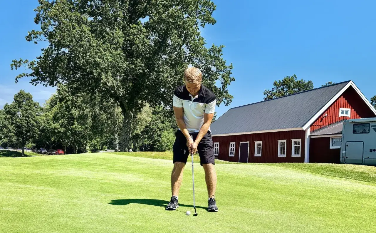Man puttar p&aring; golfbana, perfekt f&ouml;r friskv&aring;rdsbidrag golf. R&ouml;d byggnad i bakgrunden.