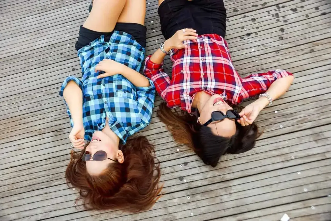 Dwie przyjaciółki leżą na drewnianej podłodze, śmiejąc się. Idealny pomysł na to, co można robić z bff w domu.