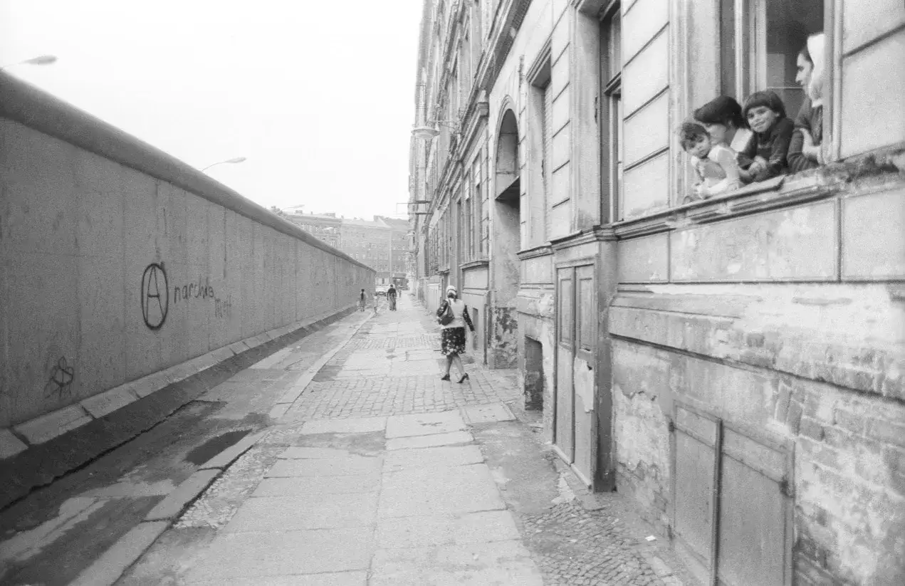 Kinder schauen neugierig aus einem Fenster auf die Berliner Mauer. Ein Anarchiesymbol ziert die Mauer. Berlin 80er Jahre.