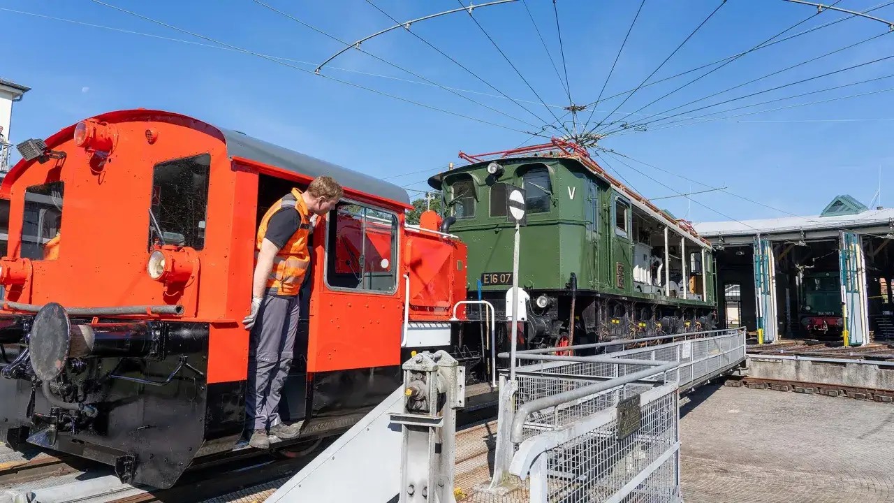 Ein Mann in Warnweste inspiziert eine rote Lokomotive neben einer gr&uuml;nen E-Lok im Eisenbahnmuseum Dieringhausen.