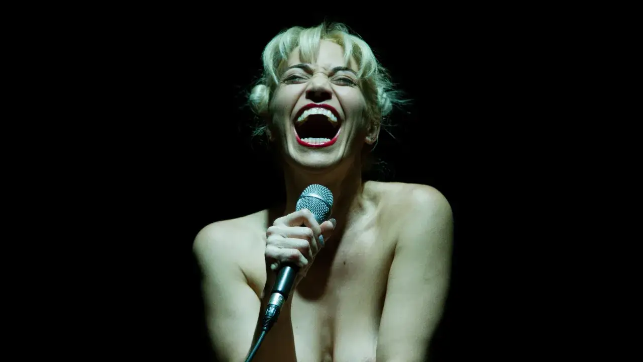 Una donna con i capelli biondi raccolti urla nel microfono, con la bocca spalancata e le labbra rosse. Sembra che stia cantando con tutta la sua anima, ma la sua espressione è così intensa che sembra quasi che stia urlando contro la merda della vita.