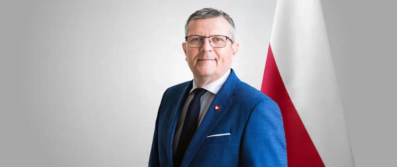 Marcin Bosacki w niebieskim garniturze, z przypinką w kształcie serca, na tle polskiej flagi.