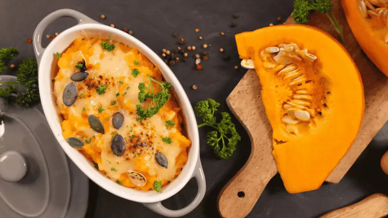 Un gratin de butternut doré et gratiné, garni de graines de courge et de persil frais. Une tranche de courge butternut crue à côté.