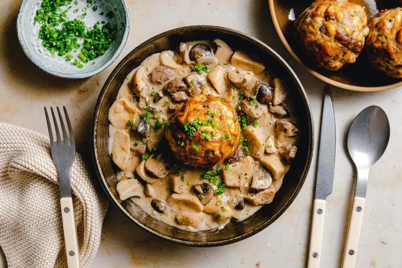 Ein herzhaftes veganes Knödel Rezept mit cremiger Pilzsoße, garniert mit frischem Schnittlauch. Dazu ein Messer und eine Gabel.