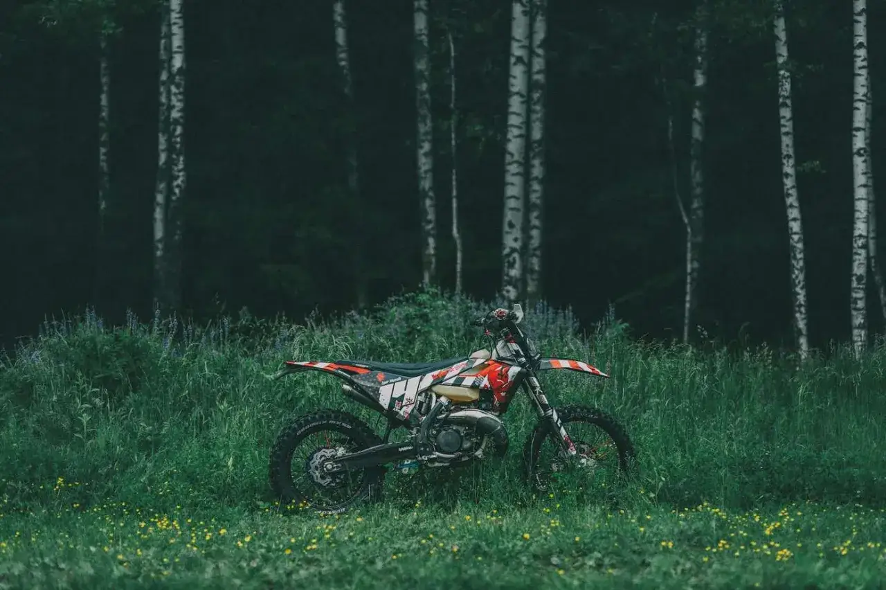 Motocykl crossowy w trawie, gotowy na przygodę. Idealne miejsce, gdzie można jeździć crossem, wśród drzew i dzikich kwiatów.