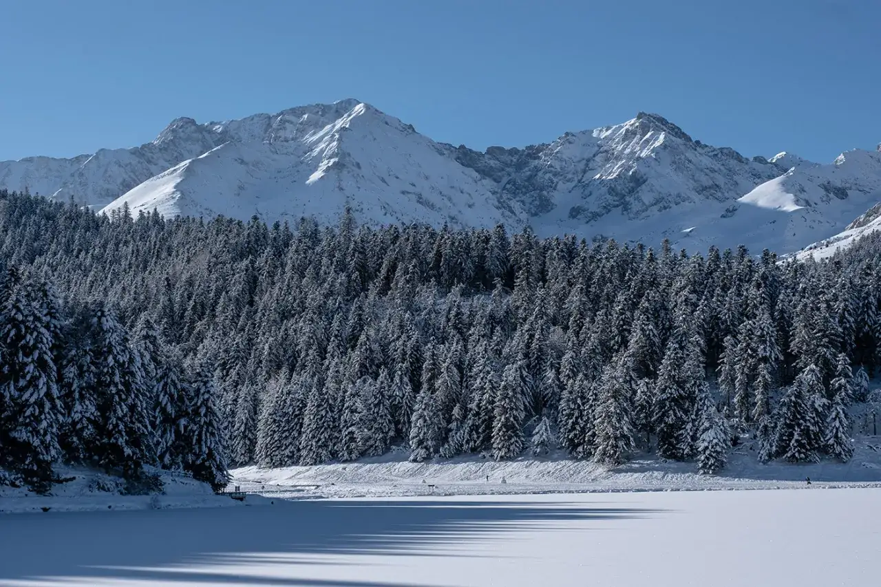 Paysage hivernal du lac de Payolle, avec des montagnes enneig&eacute;es en arri&egrave;re-plan et une for&ecirc;t de sapins recouverts de neige.