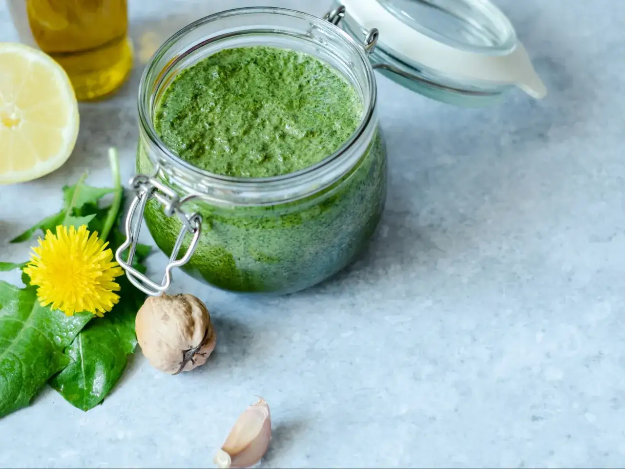 Słoik pełen zielonego pesto z liści rzodkiewki, obok cytryna, orzech, czosnek i kwiat mniszka.